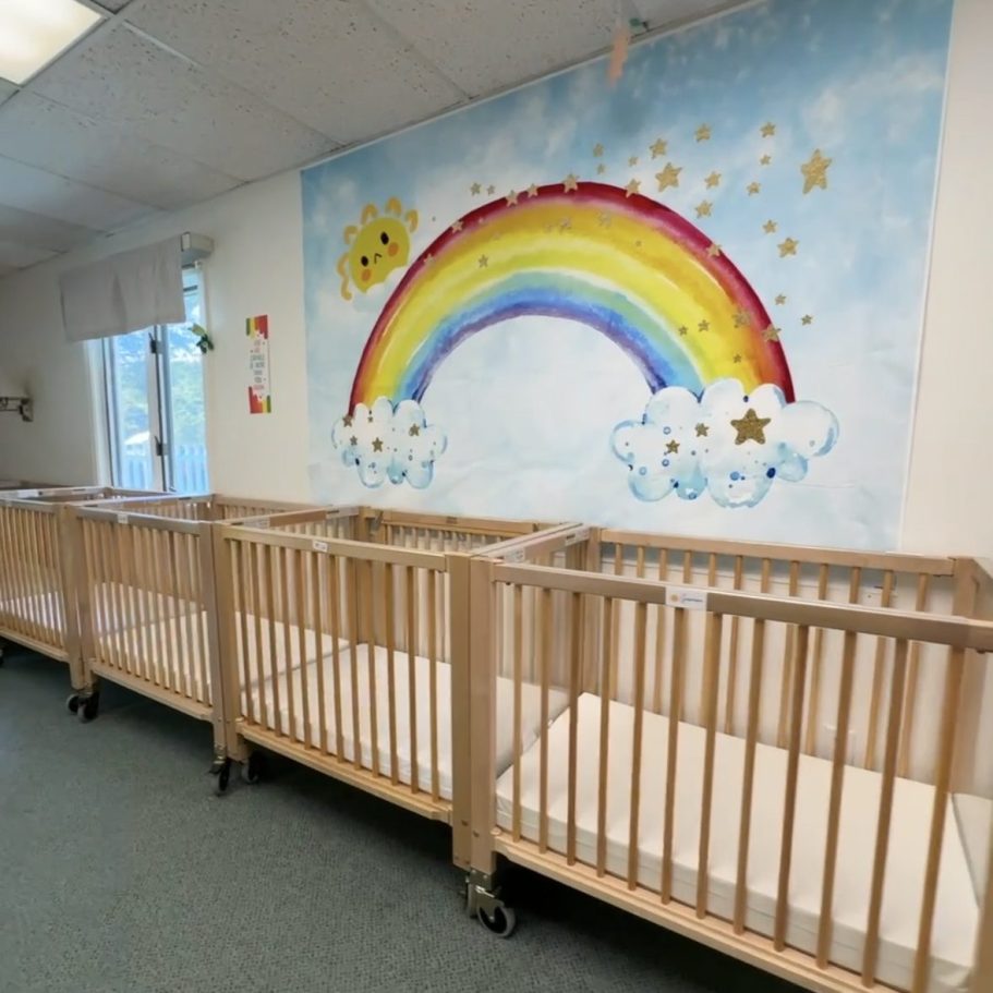 Photo displaying cribs for naps with a rainbow painting on the wall.